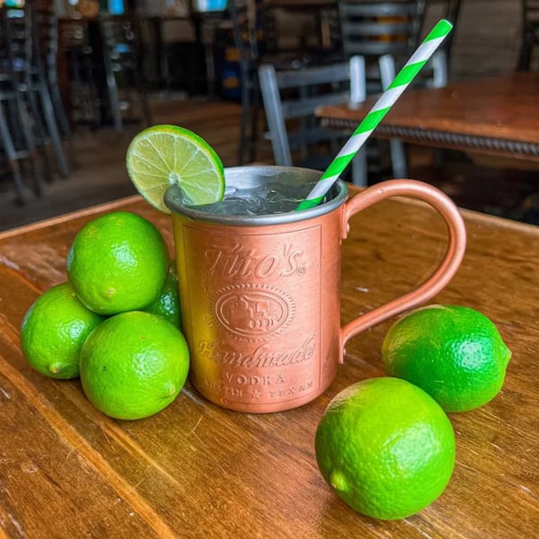 A copper Tito's Moscow Mule mug with a lime slice and striped straw, surrounded by fresh limes.
