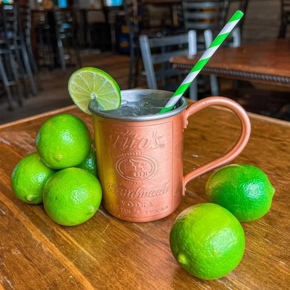 A copper Tito's Moscow Mule mug with a lime slice and striped straw, surrounded by fresh limes.