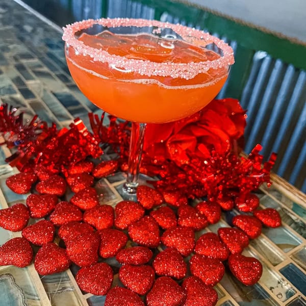 A red cocktail with a sugar-rimmed glass, surrounded by red tinsel and glitter hearts.