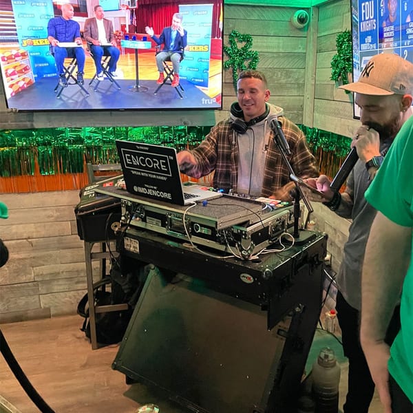A DJ with headphones and plaid shirt mixing music at a Shore House Tavern event with a TV in the background.