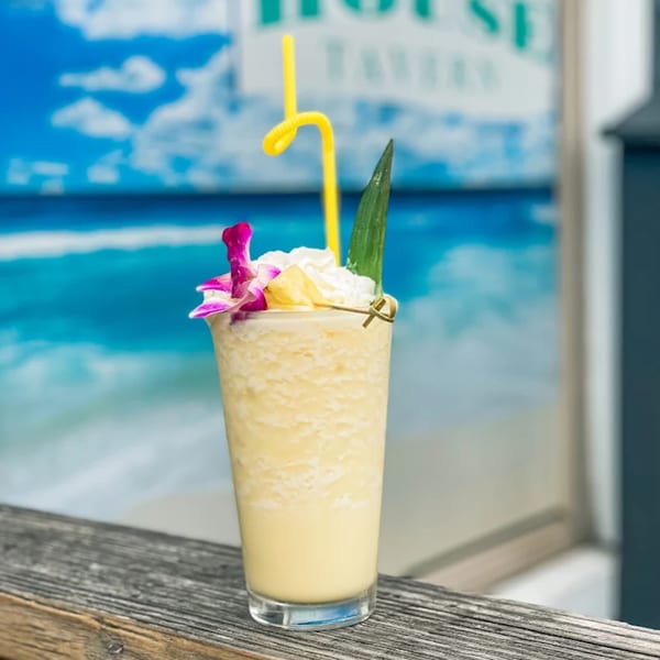 A tropical Shore House Tavern drink with whipped cream, pineapple, and an orchid, against a beach backdrop.