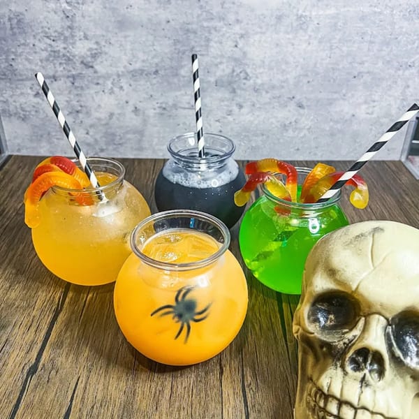 Four Halloween-themed drinks in cauldron-like glasses with striped straws and a skull decoration.