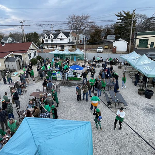 Overhead view of a lively outdoor bar-and-grill patio with many people in green attire, tents, and tables.