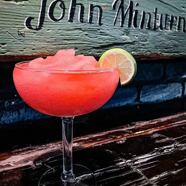 A vibrant red frozen drink with a lime wedge, set against a rustic wooden wall at The Shore House Tavern.