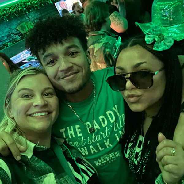 Three smiling people in green attire celebrating St. Patrick's Day at The Shore House Tavern.